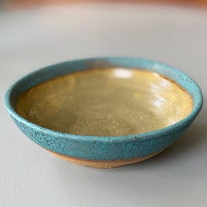 May include: A handmade ceramic bowl with a turquoise rim and a golden-yellow interior. The bowl has a rustic, textured appearance, showcasing the natural tones of the clay and glaze. The bowl is perfect for serving food.