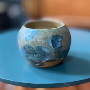 May include: A handcrafted ceramic bowl with a rounded form. The bowl has a textured surface with a mix of earthy hues, such as beige, brown, and areas of blue and teal. The inside is a warm beige colour.