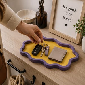 Può includere: Un vassoio giallo e viola con bordi ondulati contiene chiavi e un portafoglio. Il vassoio è appoggiato su una superficie di legno accanto a un cartello incorniciato con la scritta "It's good to be home" e una piccola pianta in vaso. Una mano raggiunge le chiavi.