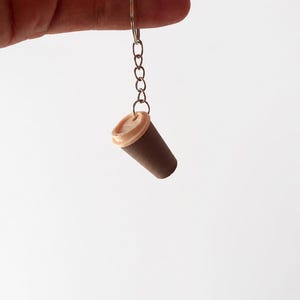 May include: A miniature coffee cup keychain featuring a dark brown cylindrical body and a light beige lid. The cup is attached to a silver-toned metal split ring by a short chain, creating a small accessory for keys or bags with a simple design.