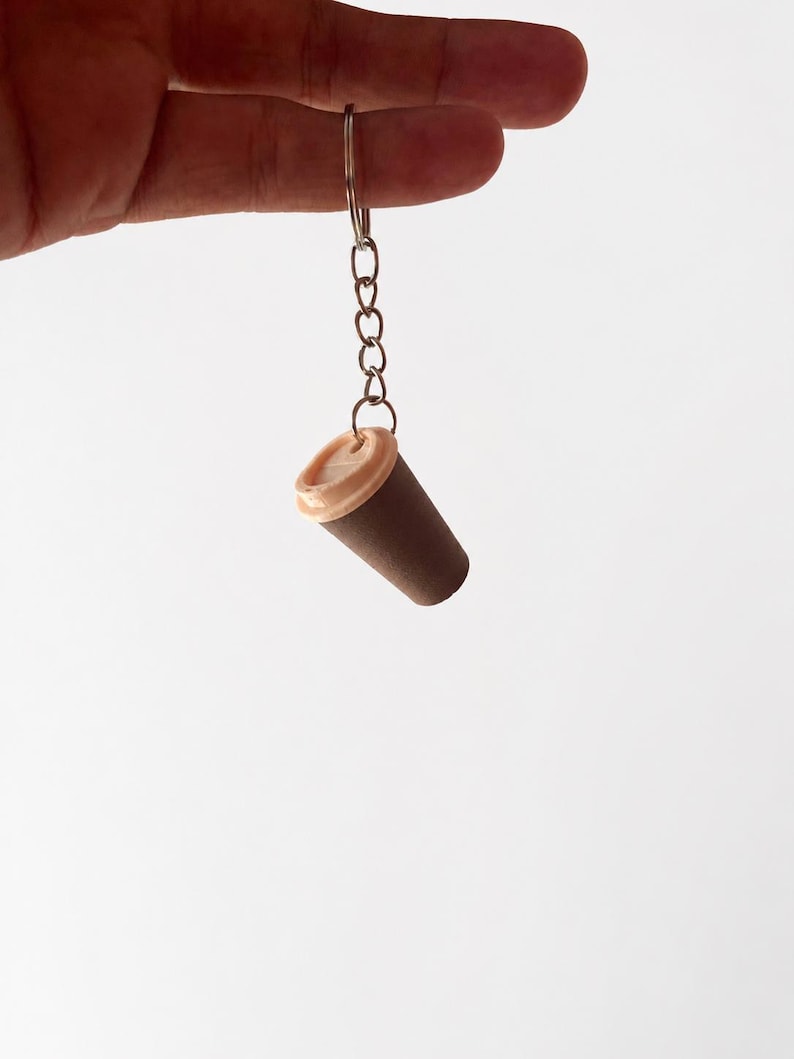 May include: A miniature coffee cup keychain featuring a dark brown cylindrical body and a light beige lid. The cup is attached to a silver-toned metal split ring by a short chain, creating a small accessory for keys or bags with a simple design.