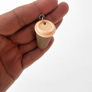 May include: A miniature coffee cup keychain featuring a light beige plastic lid and a textured, tan-colored cylindrical body. The cup is attached to a small silver-toned metal jump ring, designed for easy attachment to keys or other personal items.