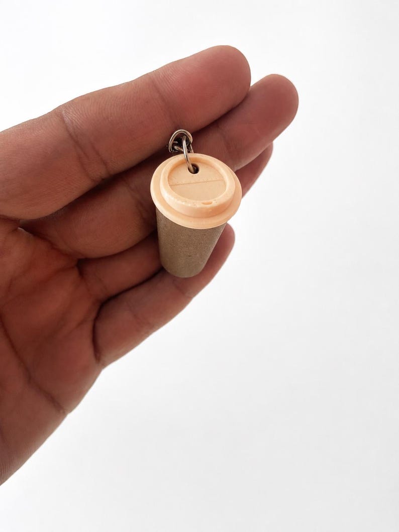 May include: A miniature coffee cup keychain featuring a light beige plastic lid and a textured, tan-colored cylindrical body. The cup is attached to a small silver-toned metal jump ring, designed for easy attachment to keys or other personal items.