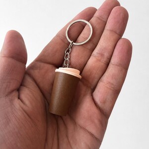 May include: A miniature coffee cup keychain featuring a dark brown cylindrical body and a light peach-colored lid. The cup is attached to a silver-toned metal split ring by a short chain, creating a compact accessory for keys or bags.