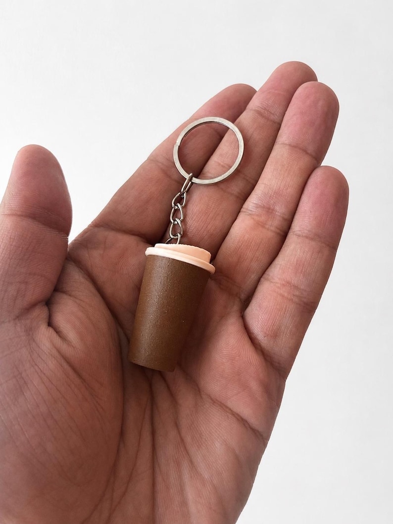 May include: A miniature coffee cup keychain featuring a dark brown cylindrical body and a light peach-colored lid. The cup is attached to a silver-toned metal split ring by a short chain, creating a compact accessory for keys or bags.