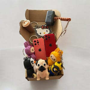 May include: A cardboard box filled with various keychains including a gray hammer, a Hello Kitty figure, two miniature red and coral phone models, a purple creature, a black and white penguin, a tan pug, a yellow creature, and a yellow minion.