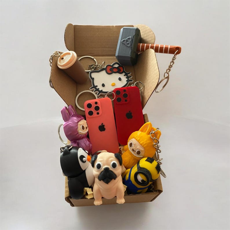 May include: A cardboard box filled with various keychains including a gray hammer, a Hello Kitty figure, two miniature red and coral phone models, a purple creature, a black and white penguin, a tan pug, a yellow creature, and a yellow minion.