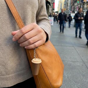 May include: A miniature coffee cup keychain featuring a dark brown base and a light beige lid, attached to a silver-toned metal chain and ring. The small, plastic-like coffee cup charm hangs from the strap of a tan leather shoulder bag.
