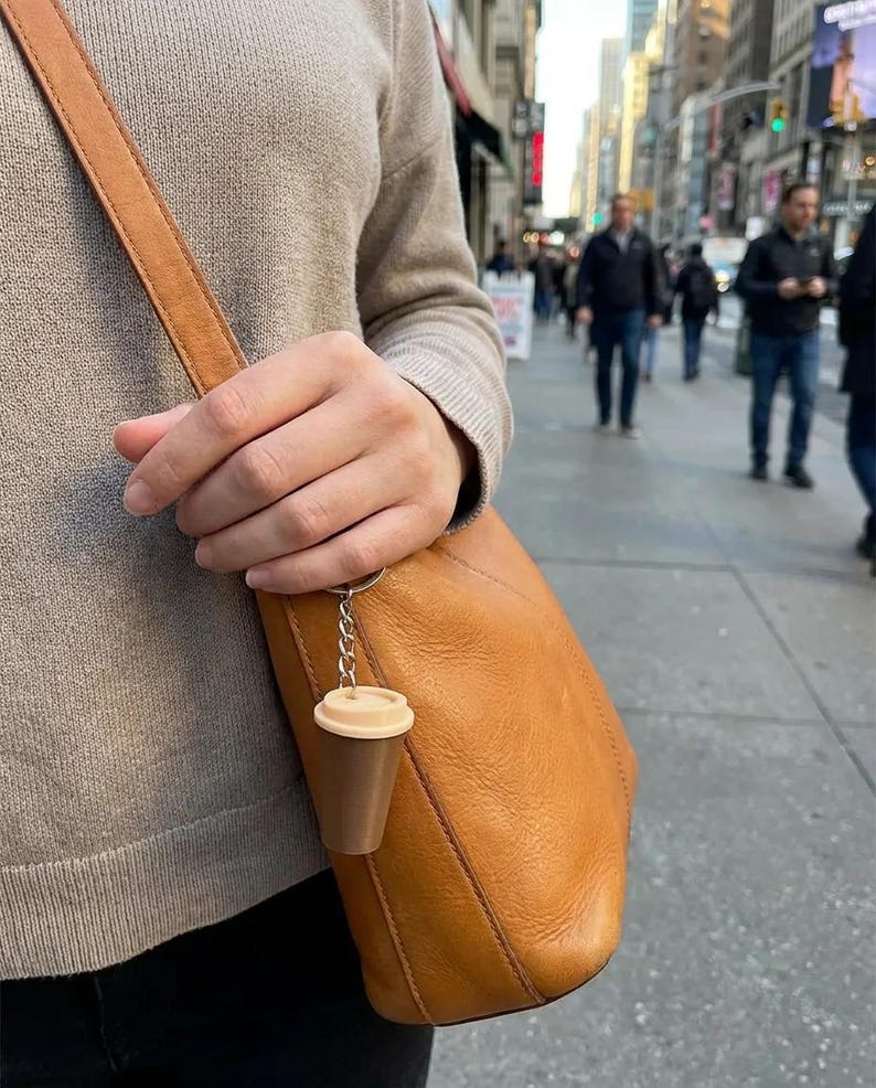 May include: A miniature coffee cup keychain featuring a dark brown base and a light beige lid, attached to a silver-toned metal chain and ring. The small, plastic-like coffee cup charm hangs from the strap of a tan leather shoulder bag.