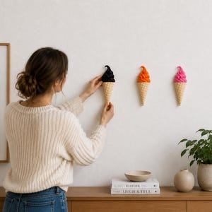 May include: Three wall-mounted ice cream cone sculptures featuring textured waffle cones and swirled tops in black, orange, and bright pink. Each piece is shaped like a soft-serve dessert and is attached to the wall in a horizontal row.