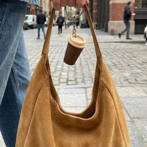 May include: A tan suede hobo bag with a silver metal keyring attached to the handle. The keyring features a miniature brown and beige coffee cup charm with a lid. The bag has a soft, slouchy structure and a single wide shoulder strap.