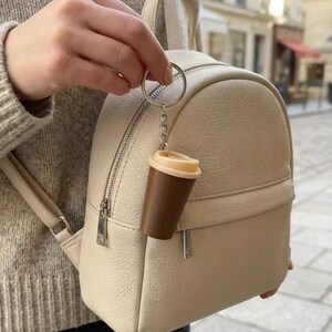 May include: A beige textured leather mini backpack featuring a silver zipper closure and a top handle. Attached to the zipper pull is a small coffee cup keychain with a dark brown body and a light beige lid, hanging from a silver metal ring.
