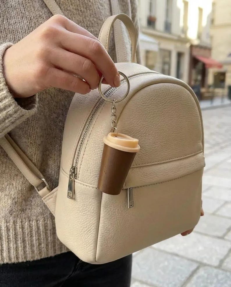 May include: A beige textured leather mini backpack featuring a silver zipper closure and a top handle. Attached to the zipper pull is a small coffee cup keychain with a dark brown body and a light beige lid, hanging from a silver metal ring.