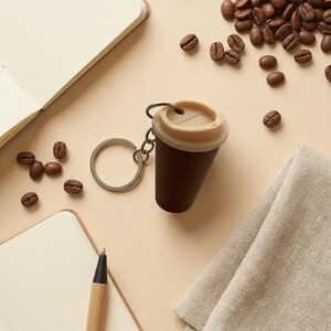 May include: A keychain shaped like a miniature dark brown coffee cup with a light beige lid. The cup features a small circular opening on the top and is attached to a silver-colored metal split ring by a short chain link connector.