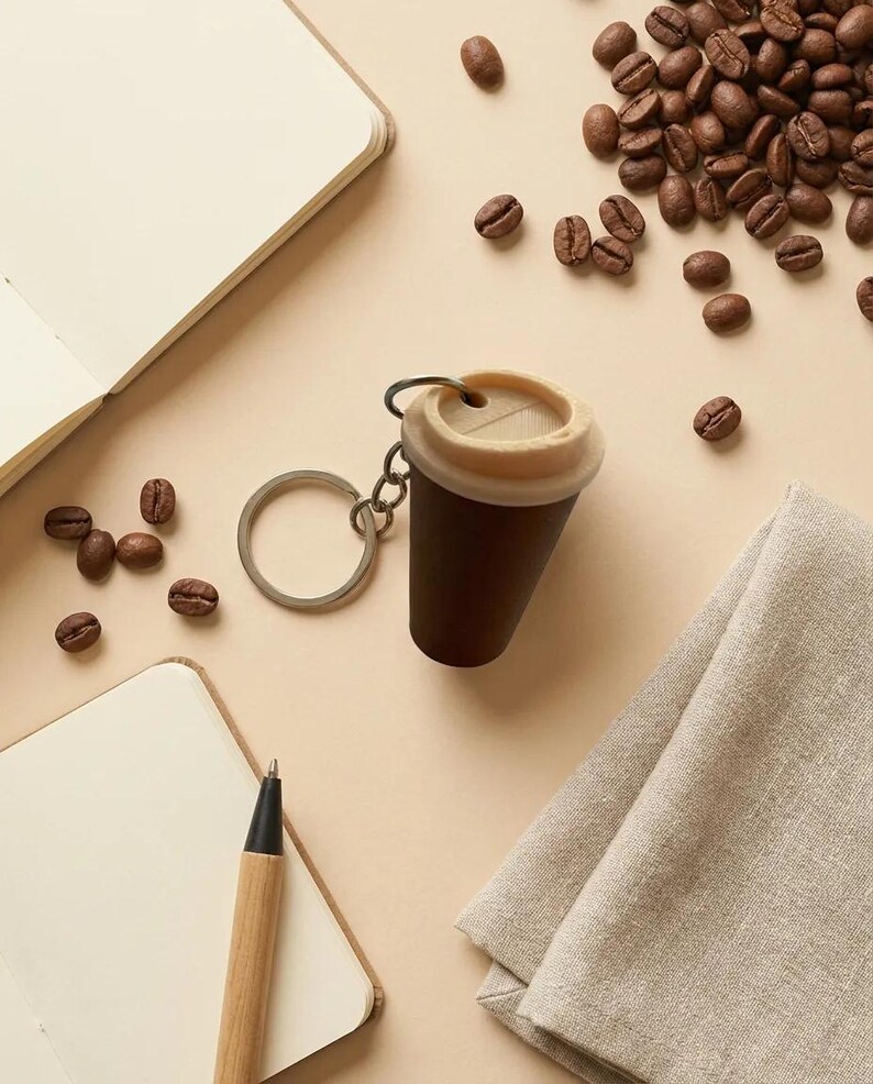May include: A keychain shaped like a miniature dark brown coffee cup with a light beige lid. The cup features a small circular opening on the top and is attached to a silver-colored metal split ring by a short chain link connector.