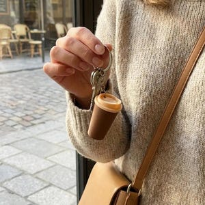 May include: A miniature coffee cup keychain featuring a dark brown cylindrical body and a light tan lid, attached to a silver metal split ring with keys. The keychain is held up, showcasing the coffee cup design in a compact, portable format.