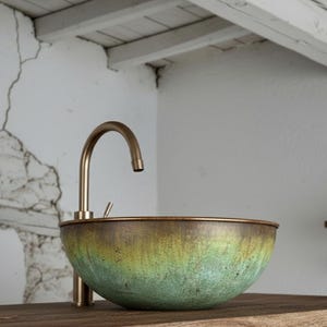 Puede incluir: Un lavabo de color cobre con un efecto degradado verde y dorado, combinado con un grifo de latón curvado. El lavabo está sobre una encimera de madera, con una pared y un techo blancos rústicos en el fondo. Dos jarras blancas están sobre una estantería de madera.