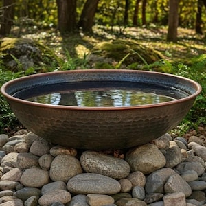 以下が含まれることがあります： 水で満たされた、大きなハンマーで叩かれた銅製のボウル。川石のベッドの上に置かれています。ボウルは暗く、質感のある外観と滑らかで反射性の高い内観を持っています。背景には緑豊かな緑と木々があります。