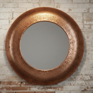 Pode incluir: Espelho redondo cor de cobre com moldura texturizada, montado numa parede de tijolos. A moldura tem um design martelado. O design geral é rústico e elegante.