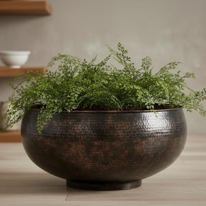 May include: A large, round, hammered copper planter filled with green foliage. The planter has a dark, aged finish and sits on a wooden surface. The background includes wooden shelves and decorative items.