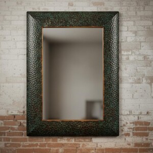 May include: Rectangular mirror with a textured, dark green frame. The frame has a hammered metal appearance with a bronze-colored inner edge. The mirror is mounted on a brick wall.