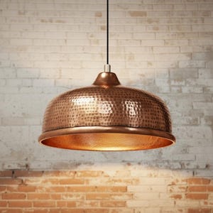 Op de afbeelding: Koperkleurige hanglamp met een gehamerde textuur. De kap in de vorm van een koepel heeft een brede rand en een gepolijst interieur. Hij hangt aan een zwart snoer. De achtergrond is een bakstenen muur.