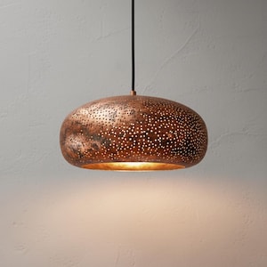 Könnte beinhalten: Eine kupferfarbene Pendelleuchte mit perforiertem Design. Die Leuchte hat eine abgerundete Form und ist an einem schwarzen Kabel aufgehängt. Die Oberfläche der Leuchte hat ein gesprenkeltes Aussehen, und die Perforationen lassen das Licht durchscheinen.