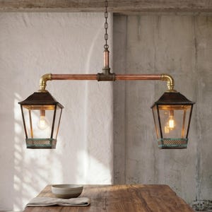 Può includere: Lampada a sospensione a due luci con struttura in rame e ottone. La lampada presenta due paralumi a forma di lanterna con lampadine a vista. L'apparecchio è appeso sopra un tavolo di legno.