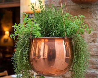 May include: A hanging copper planter filled with vibrant green herbs. The planter has a rounded shape and is suspended by copper wires. The herbs, including rosemary and other varieties, cascade over the sides, creating a lush, natural display.
