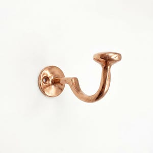May include: A polished copper wall hook featuring a circular mounting plate with a single screw hole and a curved arm that extends outward, ending in a rounded, flat-topped knob designed for hanging items securely on a vertical surface.