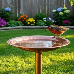 Puede incluir: Un bebedero de cobre con un pájaro pequeño posado en el borde, lleno de agua. El pájaro tiene el pecho rojo y plumas marrones. El bebedero está en un jardín con hierba verde y flores coloridas.