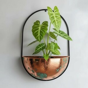 Op de afbeelding: Een aan de muur gemonteerde plantenbak met een koperkleurige, gehamerde metalen kom en een zwart metalen frame. De plantenbak bevat een groene plant met grote, geaderde bladeren. De plantenbak is ongeveer 30 cm breed en 46 cm hoog.