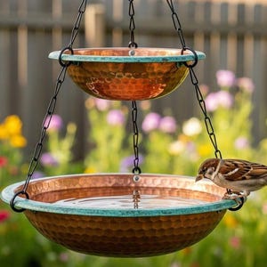 Pode incluir: Um bebedouro para pássaros de cobre de dois níveis com borda turquesa, suspenso por correntes pretas. Um pequeno pássaro está empoleirado na tigela inferior, bebendo água. O fundo apresenta um jardim desfocado com flores coloridas e uma cerca de madeira.