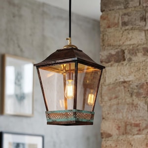 May include: A vintage-style pendant light fixture with a bronze-colored metal frame and clear glass panels. The light has two Edison-style bulbs and a decorative, aged-copper base. The fixture hangs from a black cord and is mounted near a brick wall.