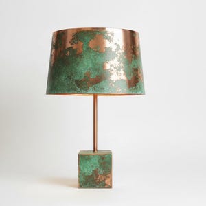 May include: A copper table lamp with a square base and a cylindrical shade. The shade and base have a green patina, while the stem is copper-coloured. The lamp is a decorative lighting fixture.