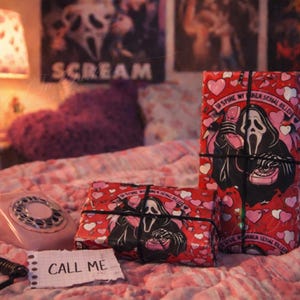 Può includere: Due regali confezionati con un motivo a cuori rossi e una grafica nera di un fantasma che tiene in mano un telefono. I regali sono legati con spago nero. Un telefono a disco rosa e un biglietto con "CALL ME" sono anche nell'immagine.