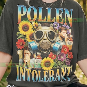 May include: Dark gray t-shirt with a colorful graphic. The design features the word "POLLEN" above "INTOLERANT", a gas mask, sunflowers, and people sneezing. The shirt also includes bottles of allergy relief.