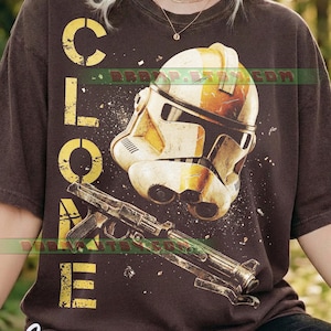 May include: Dark brown t-shirt featuring a distressed design with the word "CLONE" in yellow block letters. The shirt displays a detailed illustration of a Clone Trooper helmet and a blaster rifle, with the word "Espresso" in cursive.