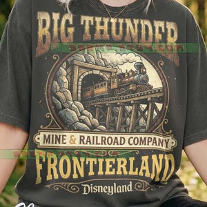 May include: Dark grey t-shirt with a vintage train graphic, featuring the text "BIG THUNDER" and "FRONTIERLAND Disneyland". The design includes a mine entrance and the words "MINE & RAILROAD COMPANY". The shirt is a classic fit.