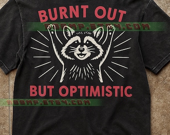Op de afbeelding: Donkergrijs t-shirt met een witte wasbeer-afbeelding met opgeheven armen en de tekst "BURNT OUT BUT OPTIMISTIC" in het rood. Het ontwerp is geplaatst tegen stralende lijnen. Het t-shirt heeft een vintage, versleten uitstraling.