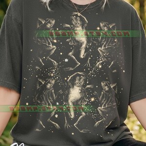 May include: Dark gray t-shirt featuring a whimsical illustration of dancing frogs surrounded by sparkling stars. The design is in a vintage style, with the word "Pepper" in a cursive font at the bottom.