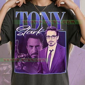 May include: Dark gray t-shirt featuring a graphic of Tony Stark. The design includes the name "TONY" in blue and the name "Stark" in white script. The image of Tony Stark is in a purple frame.