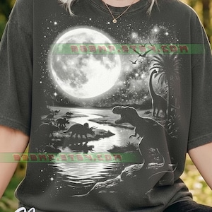 Peut inclure: T-shirt gris foncé avec une illustration en noir et blanc de dinosaures sous une pleine lune et un ciel étoilé. Le mot "Pepper" est écrit en cursive en bas du t-shirt.