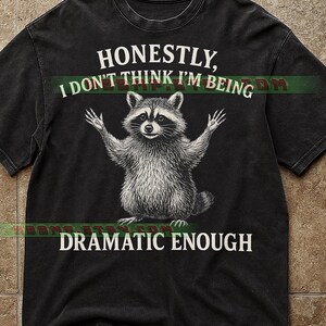 May include: Black t-shirt featuring a raccoon illustration with arms raised. The text reads "Honestly, I don't think I'm being dramatic enough." The design is in white, contrasting with the dark fabric. The shirt has a vintage, worn look.