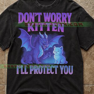 Può includere: T-shirt nero con una grafica raffigurante un drago viola e un gattino blu. Il testo "DON'T WORRY KITTEN I'LL PROTECT YOU" è stampato sopra e sotto l'illustrazione. La maglietta ha un aspetto vintage.