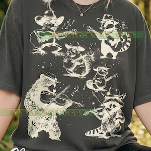 Könnte beinhalten: Dunkelgraues T-Shirt mit einer skurrilen Illustration einer Band von Tieren, darunter ein Frosch, ein Waschbär, ein Eichhörnchen und ein Bär, die alle Cowboyhüte tragen und Musikinstrumente spielen. Das Wort "Pepper" ist unten in weißer Schrift gedruckt.