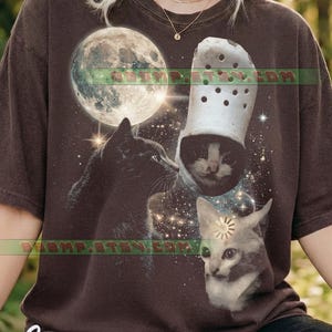 Può includere: T-shirt color espresso con un design stravagante. Presenta una luna piena, un gatto nero, un gatto in una scarpa zoccolo bianca e un gatto con un sole sulla fronte, su uno sfondo spaziale stellato.