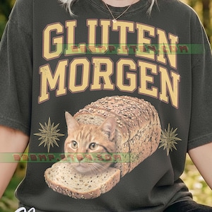 May include: A charcoal gray t-shirt featuring the text "GLUTEN MORGEN" in yellow block letters. Below the text is a graphic of an orange tabby cat head emerging from a loaf of seeded bread, flanked by two gold starburst icons on each side.