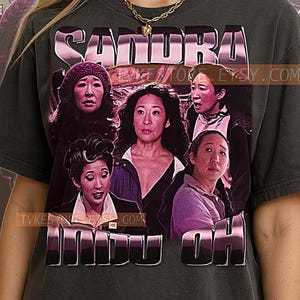 May include: Dark gray t-shirt featuring a graphic of Sandra Oh with the text "SANDRA" in a pink, metallic font above and "INDU OH" below. The graphic includes multiple images of the actress. The shirt has a crew neck and short sleeves.