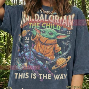 May include: Dark blue denim t-shirt with a graphic of The Mandalorian and Grogu from Star Wars. The design includes the text "The Mandalorian The Child" and "This is the Way" with planets and stars.
